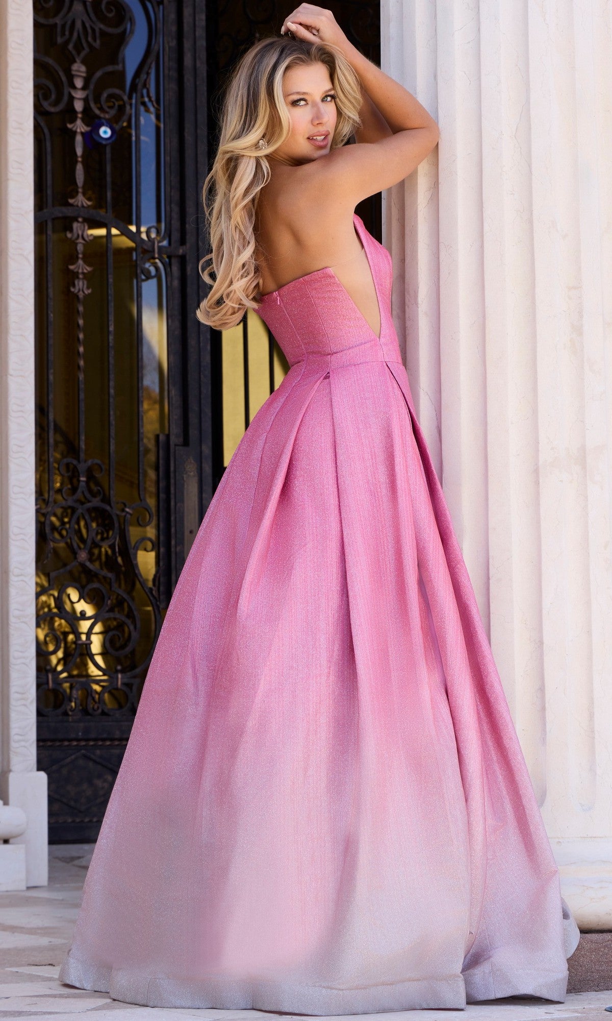 A woman wearing the Jovani 44435 long prom dress, a strapless pink ombre shimmer ball gown, stands before ornate doors and white columns, glancing back over her shoulder.